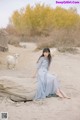 A woman in a blue dress sitting on a log in the sand.